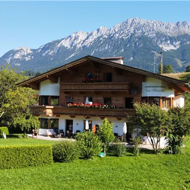 Ein traditionelles alpenländisches Haus umgeben von grünen Wiesen. Im Hintergrund sind majestätische Berge zu sehen.