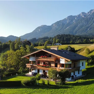 Ein schönes alpenländisches Haus mit Balkon, umgeben von grünen Wiesen. Im Hintergrund sind majestätische Berge zu sehen.