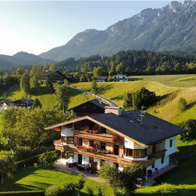 Ein malerisches Haus inmitten einer grünen Landschaft. Im Hintergrund erheben sich majestätische Berge unter einem klaren Himmel.