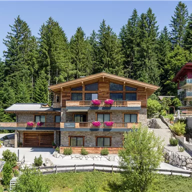 A modern chalet surrounded by green trees. The facade is adorned with flowers, and the architecture displays alpine elegance.