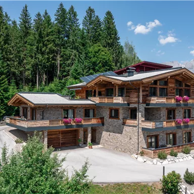 A beautiful, modern chalet made of wood and stone, surrounded by trees. The balconies are decorated with blooming plants, and the mountains are visible in the background.