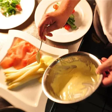 A chef is preparing a dish. On the plate are white asparagus spears and smoked salmon, next to it a bowl of sauce.