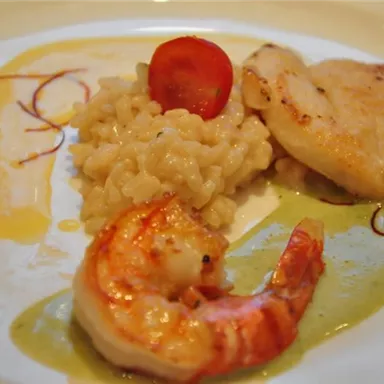 An artfully arranged plate with risotto, fried fish, and shrimp. Various sauces and a cherry tomato are decoratively placed alongside.