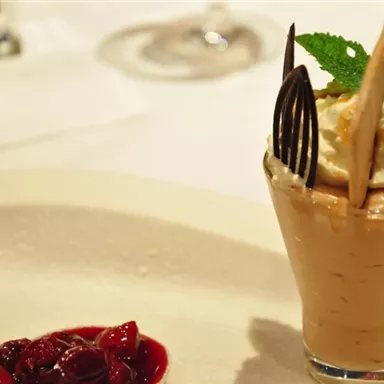 A dessert in a glass with cream, chocolate chips, and a mint leaf. Next to it lies a sauce made from red fruits on a white plate.