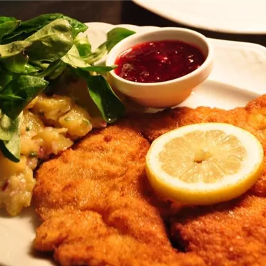 A delicious dish with breaded meat, served with a slice of lemon, potato salad, and a cup of cranberry jam. Fresh leaf salad complements the meal.