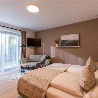 A cozy bedroom with a double bed and a small seating area. Large windows provide a view of a garden and let in plenty of daylight.