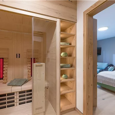 A modern sauna with a glass door and wooden shelves. In the background, a cozy bedroom can be seen.