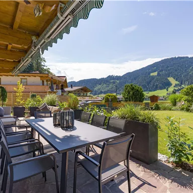 A beautiful outdoor area with a large dining table and modern chairs. In the background, green mountains and a sunny landscape can be seen.