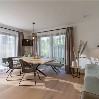 A modern living room with a dining area and large windows. The space is bright and friendly, featuring stylish furniture and decorative elements.