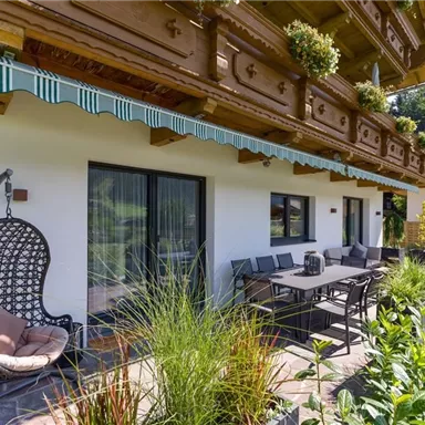 A cozy outdoor area with a large table and modern chairs. In the background, there is a hanging chair and a green garden landscape.