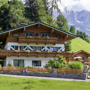 A charming wooden house surrounded by green meadows and trees. In the background, majestic mountains can be seen.