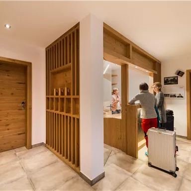 A modern reception area with a bright interior and wooden paneling. Two people are standing at a reception desk, while luggage is nearby.
