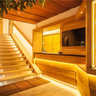 Der Empfangsbereich eines modernen Gebäudes mit Holzverkleidung und sanfter Beleuchtung. Eine Treppe führt nach oben, und es gibt eine TV-Anlage sowie Regale mit dekorativen Objekten.
