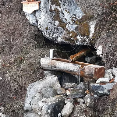 A small niche in the rocks with a wooden cross and a stream of water. Surrounded by stones and wooden logs.