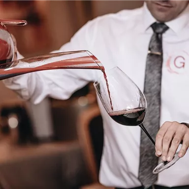 Ein Kellner gießt sorgfältig Rotwein aus einer Karaffe in ein Glas. Der Hintergrund zeigt eine elegante Restaurantatmosphäre.
