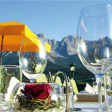 Ein eleganter Tisch mit Glaswaren und einer roten Rose in einer Vase. Im Hintergrund sind Berge und ein blauer Himmel zu sehen.