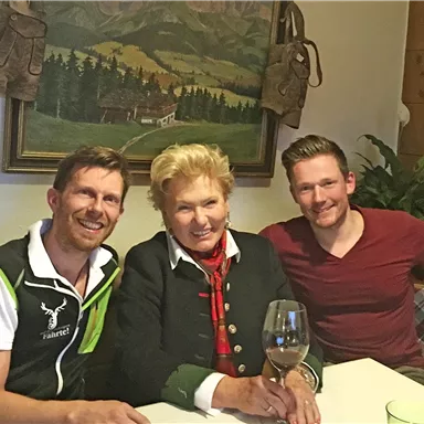 Drei Personen sitzen am Tisch in einer gemütlichen Umgebung. Eine Person hält ein Glas Wein und im Hintergrund ist ein Bild einer Landschaft zu sehen.