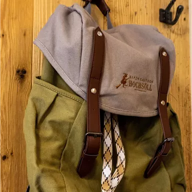 Ein grüner Rucksack mit grauem Deckel und braunen Gurten hängt an einem Haken. Der Rucksack hat eine einfache, rustikale Ausstrahlung.