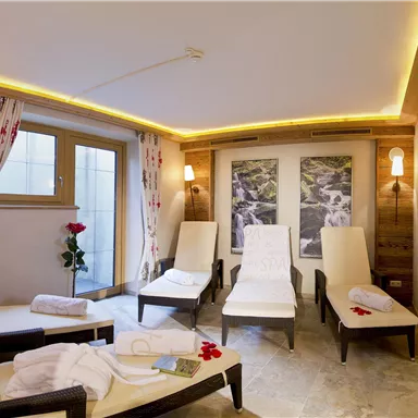 Ein entspannender Wellnessraum mit Liegen und sanfter Beleuchtung. Durch die Fenster fällt viel Tageslicht und es gibt dekorative Elemente wie Blumen.