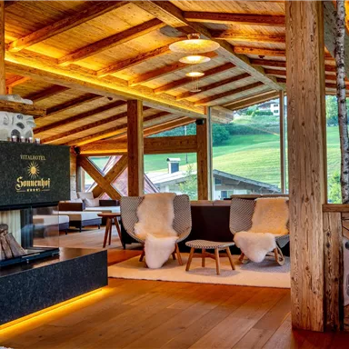 Ein gemütlicher Wohnraum mit Holzdecke und großen Fenstern. Der Kamin und die bequemen Möbel schaffen eine einladende Atmosphäre.