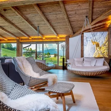 Ein modernes Wohnzimmer mit Holzdecke und großen Fenstern, die einen Blick auf die Natur ermöglichen. Bequeme Sessel und eine hängende Liege sorgen für eine gemütliche Atmosphäre.