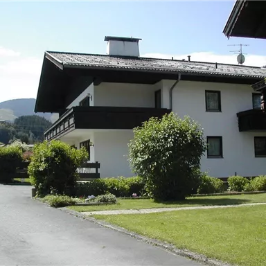 Ein ruhiges Gebäude in ländlicher Umgebung mit einem gepflegten Garten. Im Hintergrund sind sanfte Hügel und ein blauer Himmel zu sehen.