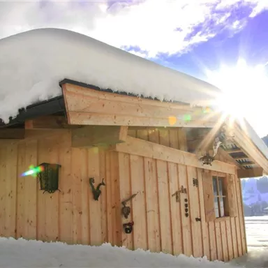 Eine gemütliche Holzhuette im Schnee mit Sonne, die hinter ihr aufgeht. Der Dachfirst ist mit einer dicken Schneeschicht bedeckt.