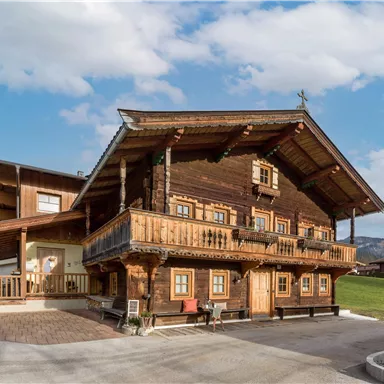 A traditional wooden house with a beautiful balcony and a bright facade. In the background, gentle hills and a blue sky can be seen.