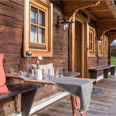 A cozy wooden house with large windows and an inviting porch. On a table, there are glasses and a vase with flowers, creating a friendly atmosphere.