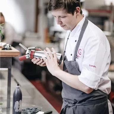 Ein Koch in einer modernen Küche bereitet ein Gericht mit einem Siphon vor. Auf dem Tisch steht ein Dessert, und im Hintergrund sind Küchengeräte zu sehen.