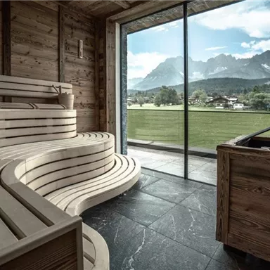 Ein modernes Saunainneres mit Holzbank und großen Fenstern. Der Blick zeigt eine malerische Landschaft mit Bergen und Wiesen.