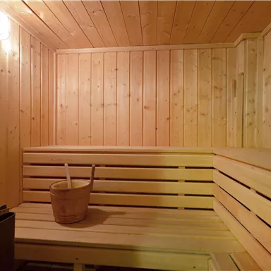 Eine gemütliche Sauna mit Holzverkleidung und Bänken. In der Ecke steht ein Holzbehälter und eine Uhr an der Wand.