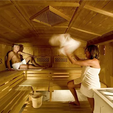 A sauna with wooden panels, where two people are relaxing. One person is swinging a towel while the other is sitting.