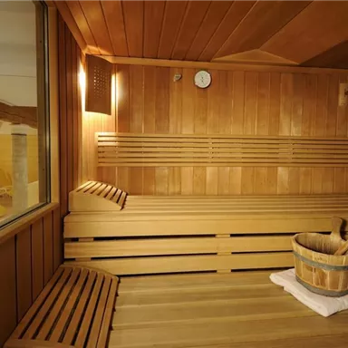 Ein gemütlicher Wellnessbereich mit Holzbank und einem Holzeimer. Die Wände sind aus hellem Holz und es gibt einen großen Fensterausschnitt.