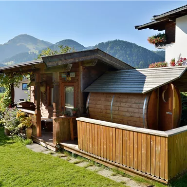 A typical wooden house with a sauna in the garden, surrounded by green grass and mountains in the background. The sky is clear and sunny.