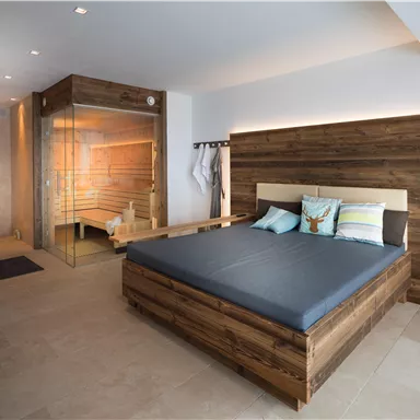 Ein modernes Schlafzimmer mit einem großen Bett und gemütlichen Kissen. Im Hintergrund befindet sich eine Sauna mit warmem Licht.