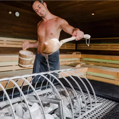 A man in a sauna pouring water on the hot stones. The surroundings are made of wood with benches for sitting.