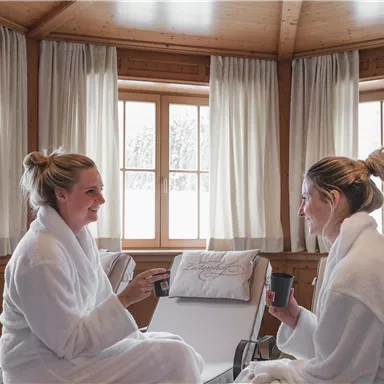 Zwei Frauen in Bademänteln entspannen in einem hellen Raum mit großen Fenstern. Sie unterhalten sich bei einer Tasse Tee in einer ruhigen, gemütlichen Atmosphäre.