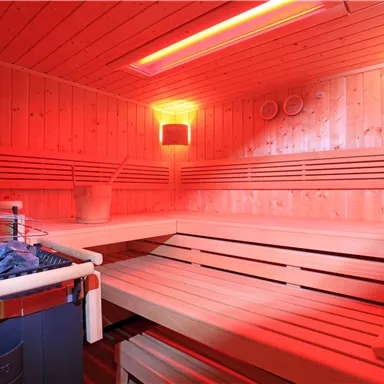 Eine moderne Sauna mit Holzverkleidung und rotem Licht. Im Vordergrund sind Steine und ein Holzbehälter zu sehen.