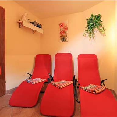 Ein entspannender Raum mit drei roten Liegen und bunten Handtüchern. An der Wand hängen ein Bild und einige Pflanzen, die eine ruhige Atmosphäre schaffen.