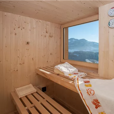 A cozy sauna room with wooden paneling and a large window view of the mountains. Towels are laid out on the benches ready for relaxation.
