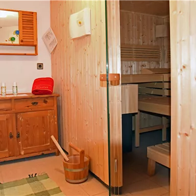 A cozy sauna area with wooden walls and a relaxation area. On the left side, there is a wooden cabinet with towels and care products.