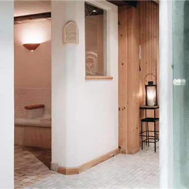 Ein entspannender Wellnessbereich mit warmen Holzwänden und sanfter Beleuchtung. Im Vordergrund ist eine Duschkabine und eine gemütliche Ecke mit einer Laterne zu sehen.