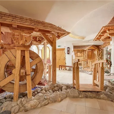 Ein rustikaler Innenbereich mit Holzstrukturen, darunter ein großes Wasserrad. Der Raum ist mit Steinen und Pflanzen geschmückt, was eine gemütliche Atmosphäre schafft.