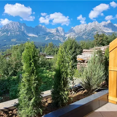 A beautiful landscape with high mountains and a blue sky. In the foreground, there are green plants and a wooden railing.