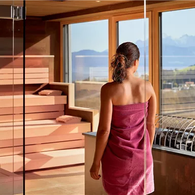 A woman in a towel stands in a modern spa. The view out of the window shows an impressive mountain landscape.