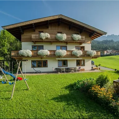 A charming house with a balcony and flower boxes, surrounded by green lawn and trees. In the foreground, there is a playground with swings.