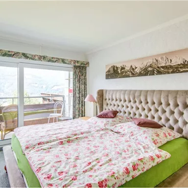 A cozy bedroom with a large bed and floral bedding. The windows offer a beautiful view of the mountains and the terrace.