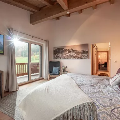 Ein gemütliches Schlafzimmer mit einem großen Bett und Holzdecken. Eine Tür öffnet sich zu einem Balkon mit Blick auf die Landschaft.
