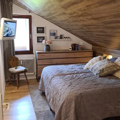 A cozy bedroom in the attic with a double bed, a wooden wardrobe, and a small table. The walls are painted in warm tones, and a window lets natural light in.